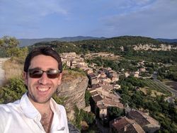 Young white man with brown hair and sunglasses wearing a white shirt in front of a village in France